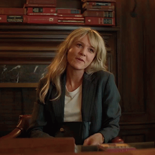 A Woman In A Suit Sits At A Desk With A Stack Of Books Behind Her That Says ' A ' On It GIF