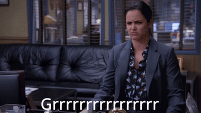 A Woman In A Suit Sits In Front Of A Couch And A Sign That Says Grrrr GIF