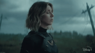 A Woman In A Superhero Costume Is Standing In A Field With A Telephone Pole In The Background . GIF