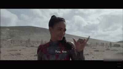 A Woman In A Superhero Costume Is Standing In The Desert And Making A Hawaiian Hand Sign . GIF