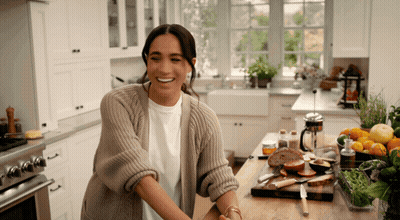 A Woman In A Sweater Is Smiling While Standing In A Kitchen GIF