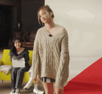 A Woman In A Sweater Is Standing In Front Of A Woman Sitting On A Yellow Couch GIF