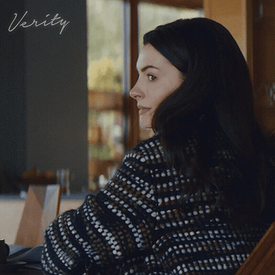 A Woman In A Sweater Sits In Front Of A Sign That Says ' Verity ' On It GIF