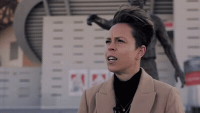 A Woman In A Tan Coat Stands In Front Of A Statue Of A Soccer Player GIF