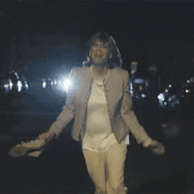 A Woman In A Tan Jacket Is Standing In The Dark GIF