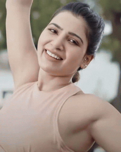 A Woman In A Tan Tank Top Smiles With Her Arm Up GIF