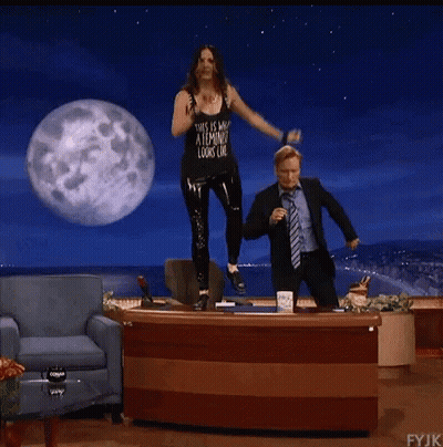 A Woman In A Tank Top That Says " This Is What A Feminist Looks Like " Stands On A Desk Next To A Man GIF