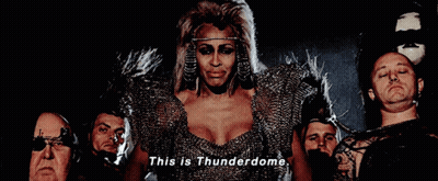 A Woman In A Thunderdome Costume Is Surrounded By Men GIF
