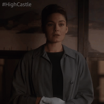 A Woman In A Trench Coat And White Gloves Stands In Front Of A Sign That Says High Castle GIF