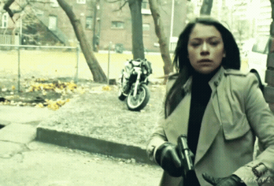 A Woman In A Trench Coat Holds A Gun In Front Of A Parked Motorcycle GIF