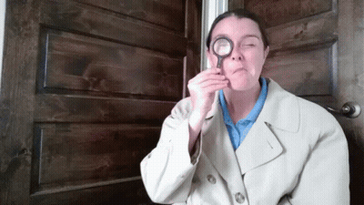 A Woman In A Trench Coat Holds A Magnifying Glass To Her Eye GIF