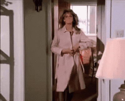 A Woman In A Trench Coat Is Standing In A Hallway . GIF