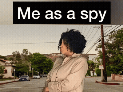 A Woman In A Trench Coat Is Standing In Front Of A Hollywood Sign GIF
