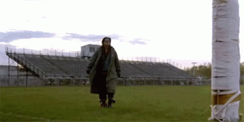 A Woman In A Trench Coat Is Walking On A Field Next To A Cigarette Butt . GIF