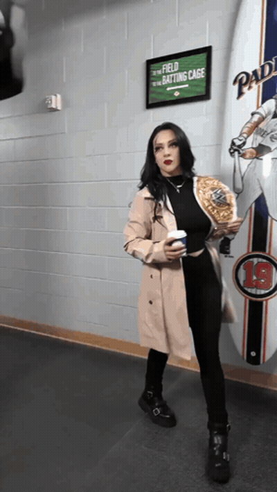 A Woman In A Trench Coat Stands In Front Of A Sign That Says " Field To The Batting Cage " GIF