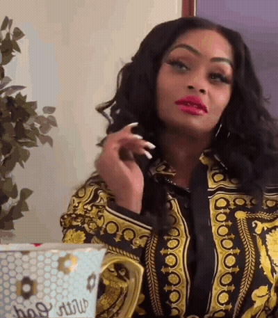 A Woman In A Versace Shirt Is Sitting At A Table With A Cup . GIF