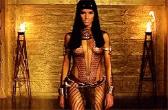 A Woman In A Very Revealing Outfit Stands In Front Of A Gold Wall GIF