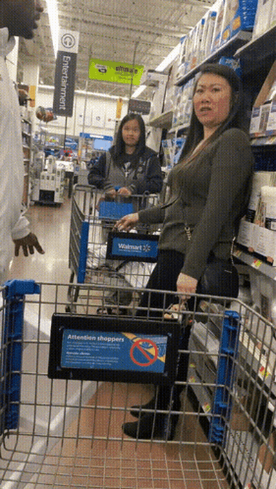 A Woman In A Walmart Shopping Cart With A Sign That Says Attention Shoppers On It GIF