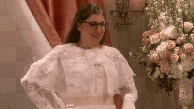 A Woman In A Wedding Dress And Gloves Is Standing Next To A Vase Of Flowers . GIF