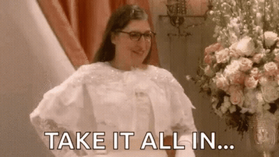 A Woman In A Wedding Dress Is Standing In Front Of A Vase Of Flowers And Says `` Take It All In '' . GIF