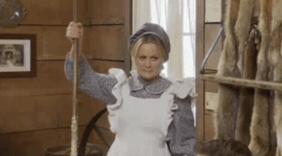 A Woman In A White Apron Is Holding A Mop In A Room . GIF