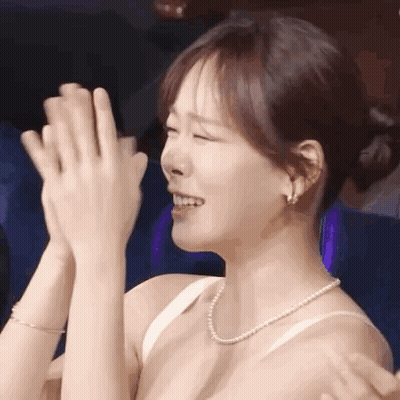 A Woman In A White Bra And Pearl Necklace Is Clapping Her Hands In A Theater . GIF
