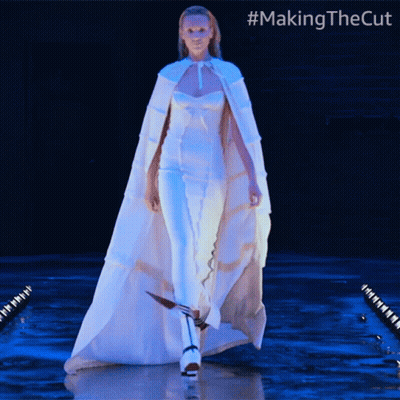 A Woman In A White Cape Is Walking Down A Runway With #Makingthecut Written On The Bottom GIF