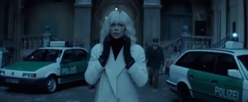 A Woman In A White Coat And Black Gloves Is Standing In Front Of A Group Of Police Cars . GIF
