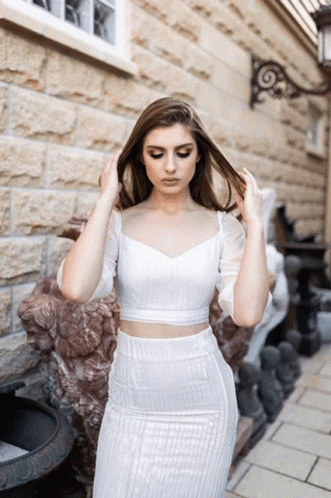 A Woman In A White Crop Top And Skirt Is Standing In Front Of A Brick Building GIF
