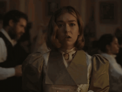 A Woman In A White Dress And A Brown Jacket Stands In A Dark Room GIF