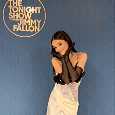 A Woman In A White Dress And Black Gloves Stands In Front Of A Sign For Jimmy Fallon GIF