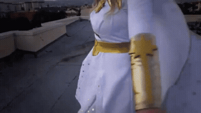 A Woman In A White Dress And Gold Gloves Is Walking On A Rooftop . GIF