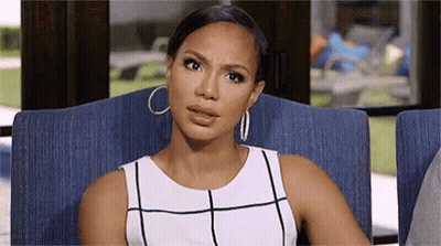 A Woman In A White Dress And Hoop Earrings Is Sitting In A Chair And Making A Funny Face . GIF