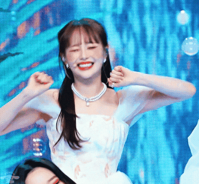A Woman In A White Dress And Pearl Necklace Is Dancing On A Stage With Her Hands In The Air . GIF