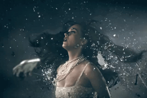A Woman In A White Dress And Pearls Is Surrounded By Pieces Of Glass GIF