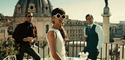 A Woman In A White Dress And Sunglasses Is Standing On A Balcony Next To Two Men . GIF