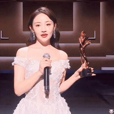 A Woman In A White Dress Holding A Microphone And A Trophy GIF