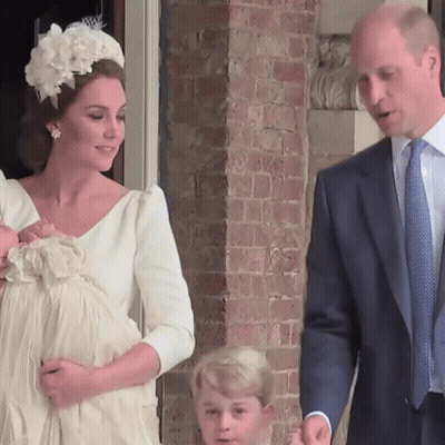 A Woman In A White Dress Holds A Baby While A Man In A Suit Looks On GIF