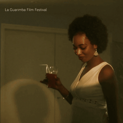 A Woman In A White Dress Holds A Glass In Front Of A La Guarimba Film Festival Poster GIF