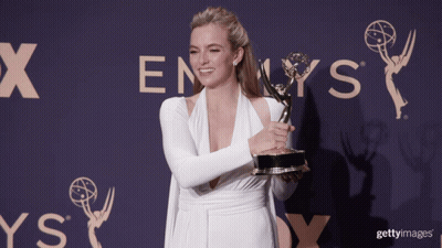 A Woman In A White Dress Holds A Trophy In Front Of An Emmys Sign GIF
