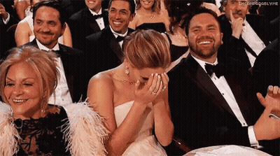A Woman In A White Dress Is Covering Her Face With Her Hands While Sitting In A Crowd Of People . GIF