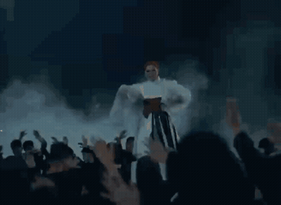 A Woman In A White Dress Is Dancing In Front Of A Crowd Of People While Flames Are Coming Out Of Her Dress . GIF