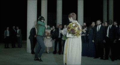 A Woman In A White Dress Is Holding A Bouquet Of Flowers In Front Of A Crowd Of People GIF