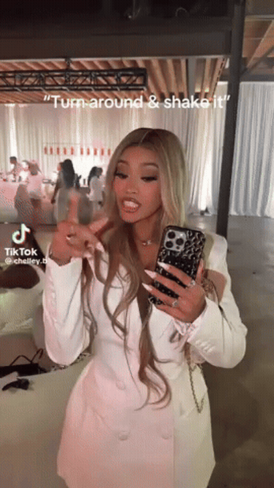 A Woman In A White Dress Is Holding A Cell Phone In Her Hand And Says " Turn Around & Shake It " GIF