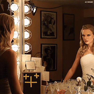 A Woman In A White Dress Is Looking At Herself In A Mirror With 4K Taylor Written On The Bottom GIF