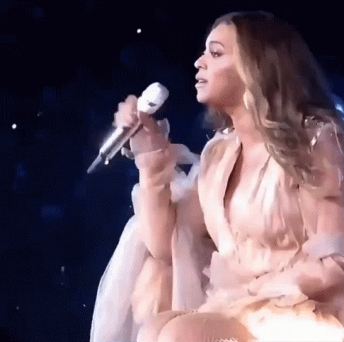 A Woman In A White Dress Is Singing Into A Microphone On Stage GIF