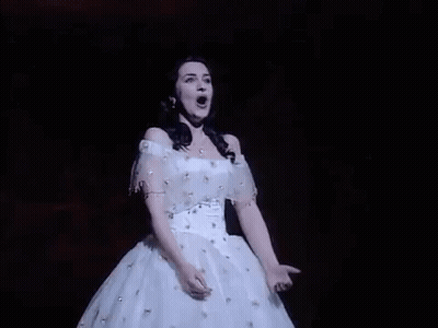 A Woman In A White Dress Is Singing On A Stage With Her Mouth Open . GIF
