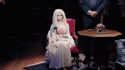 A Woman In A White Dress Is Sitting In A Chair With A Doll On Her Lap GIF