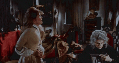 A Woman In A White Dress Is Standing In Front Of A Bed With Red Sheets GIF