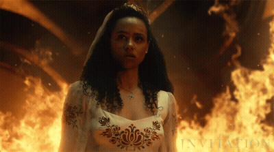 A Woman In A White Dress Is Standing In Front Of A Fire And The Word Invitation Is Visible GIF